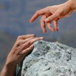 A hand reaching down to help another person climbing a rock, symbolising God helping believers when faith feels weak